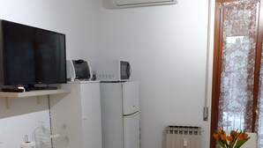 Een compacte keuken met een magnetron, broodrooster en een flatscreen televisie op een plank. Er staat een eettafel met stoelen en een vaas met bloemen.