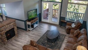 A living room with a stone fireplace, leather sofa, a television on a stand, and a large window with a view of greenery. - 3206 Golf View Hillcrest The Clubhouse (Pigeon Forge)