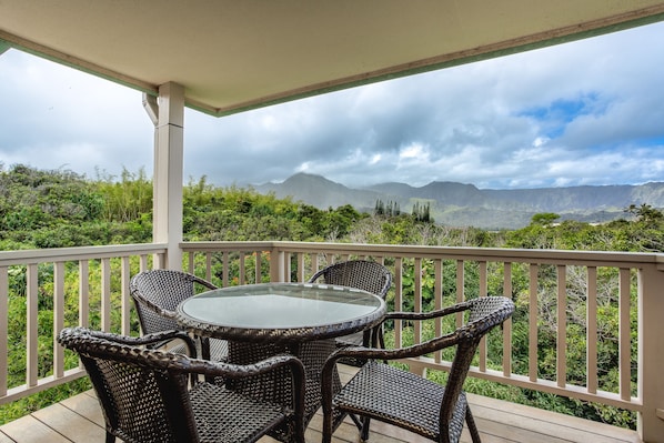 Un espacio al aire libre techado con mobiliario de mimbre y una mesa de vidrio, con vistas a un paisaje exuberante con montañas.