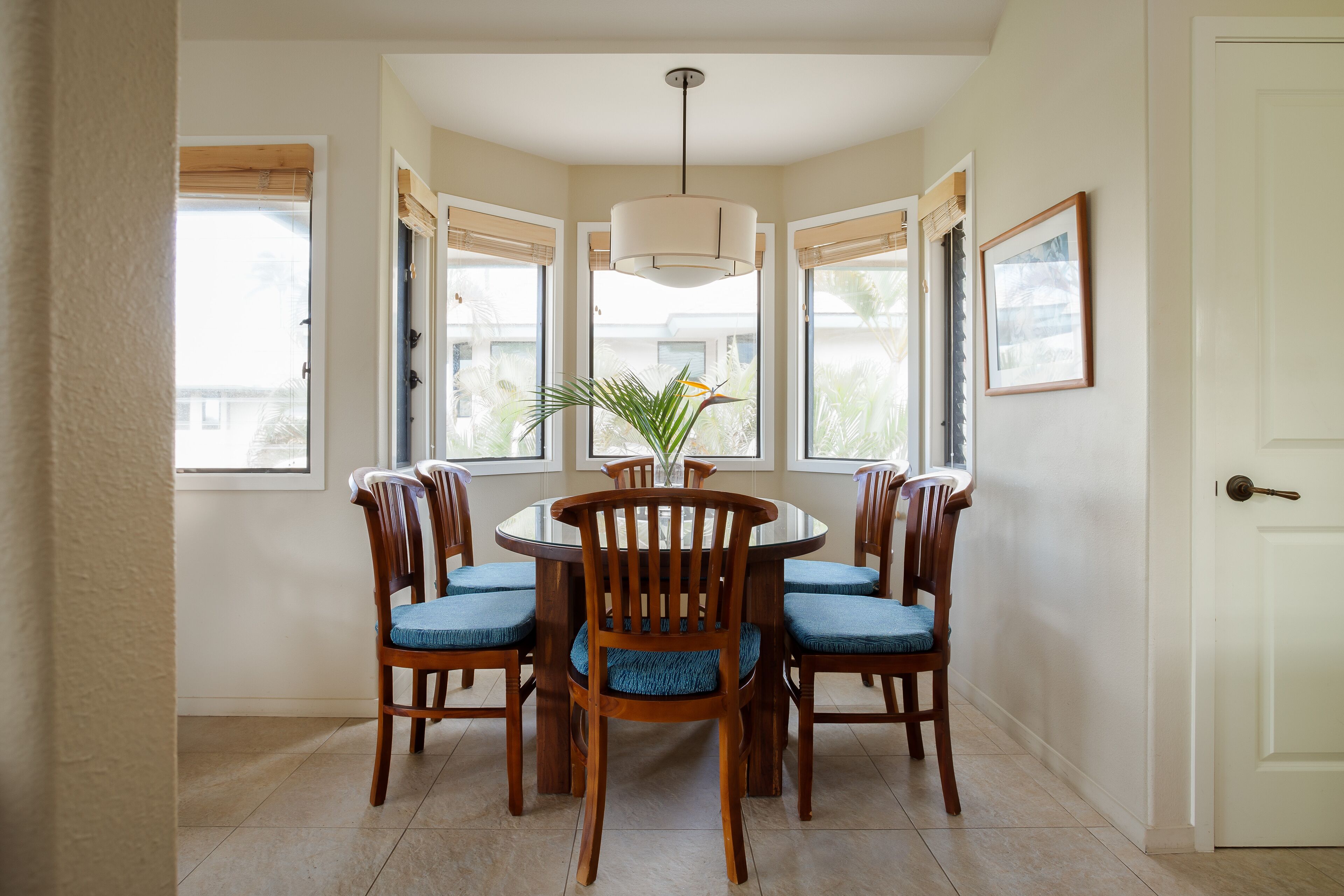 Un comedor con una mesa redonda rodeada por seis sillas de madera con cojines azules, una lámpara colgante encima y una planta en maceta sobre la mesa.