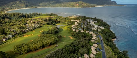 An aerial view of a coastal golf course with a road running alongside, surrounded by lush greenery and residential buildings.