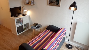 Quarto pequeno e bem iluminado, com uma cama, uma escrivaninha com computador, uma TV e uma estante com livros e uma planta.