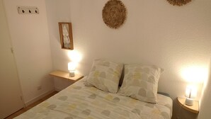Uma cama bem arrumada com roupa de cama estampada, duas luminárias de cabeceira, uma mesinha de cabeceira de madeira, um objeto decorativo fixado na parede e uma tomada na parede.