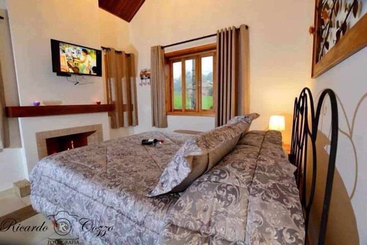 Ein Schlafzimmer mit einem großen Bett, einem an der Wand befestigten Fernseher, einem Kamin und einem Fenster mit Vorhängen.