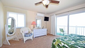 Ein Schlafzimmer mit Deckenventilator, einem Bett mit blattgemustertem Überwurf, einem Schminktisch, einem Sessel, einem Spiegel und einem an der Wand befestigten Fernseher.