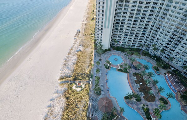Luchtbeeld van een hotel aan het strand met een groot zwembad, diverse balkons en een zandstrand.