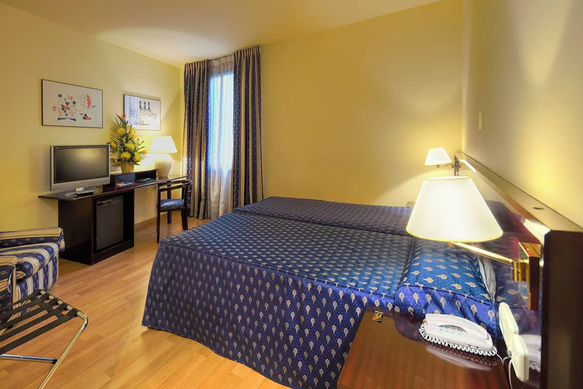 A hotel room with a bed, a desk with a computer, a TV, a chair, a lamp, and a telephone.
