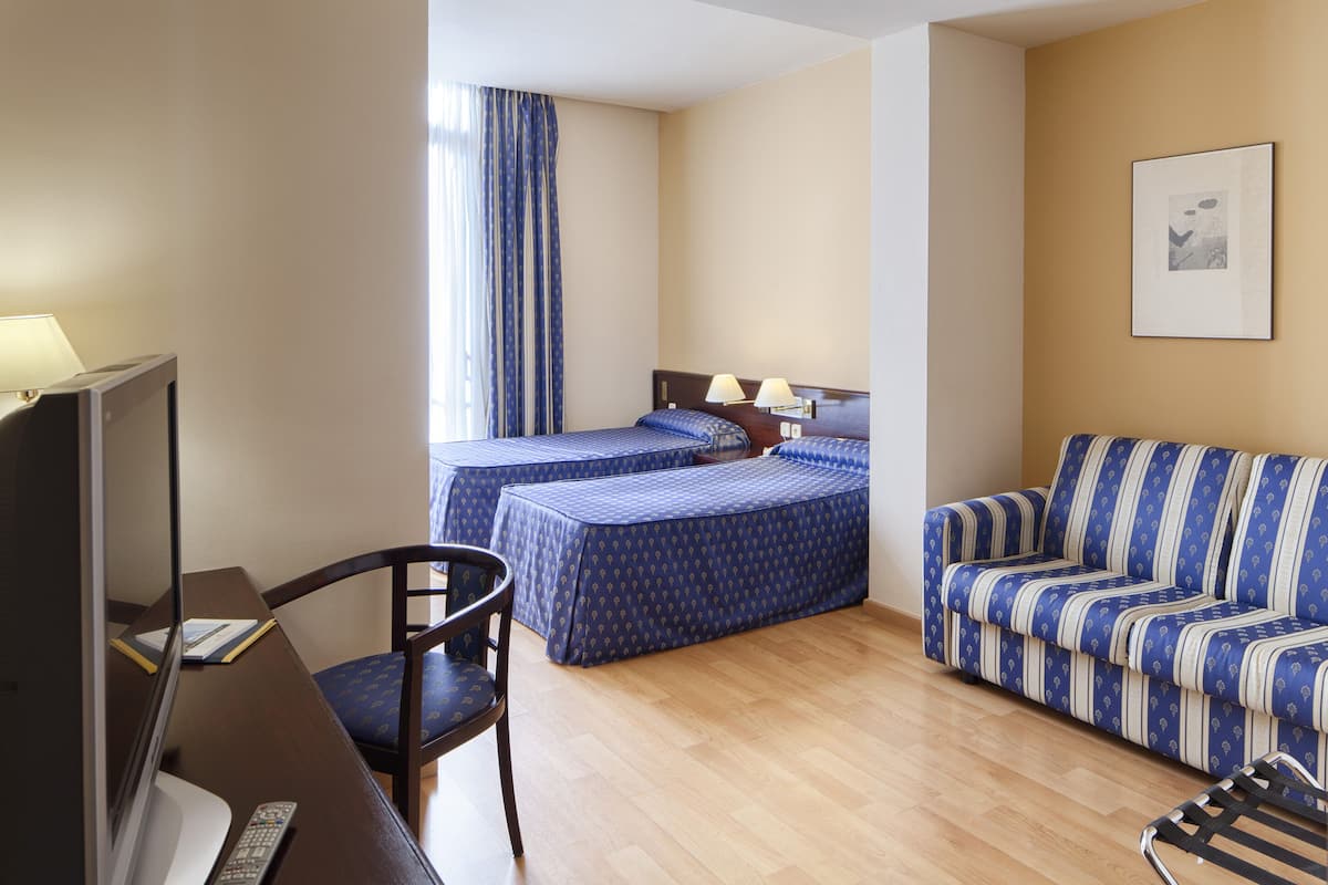 A hotel room with two beds, a sofa, a desk with a TV, and a chair.