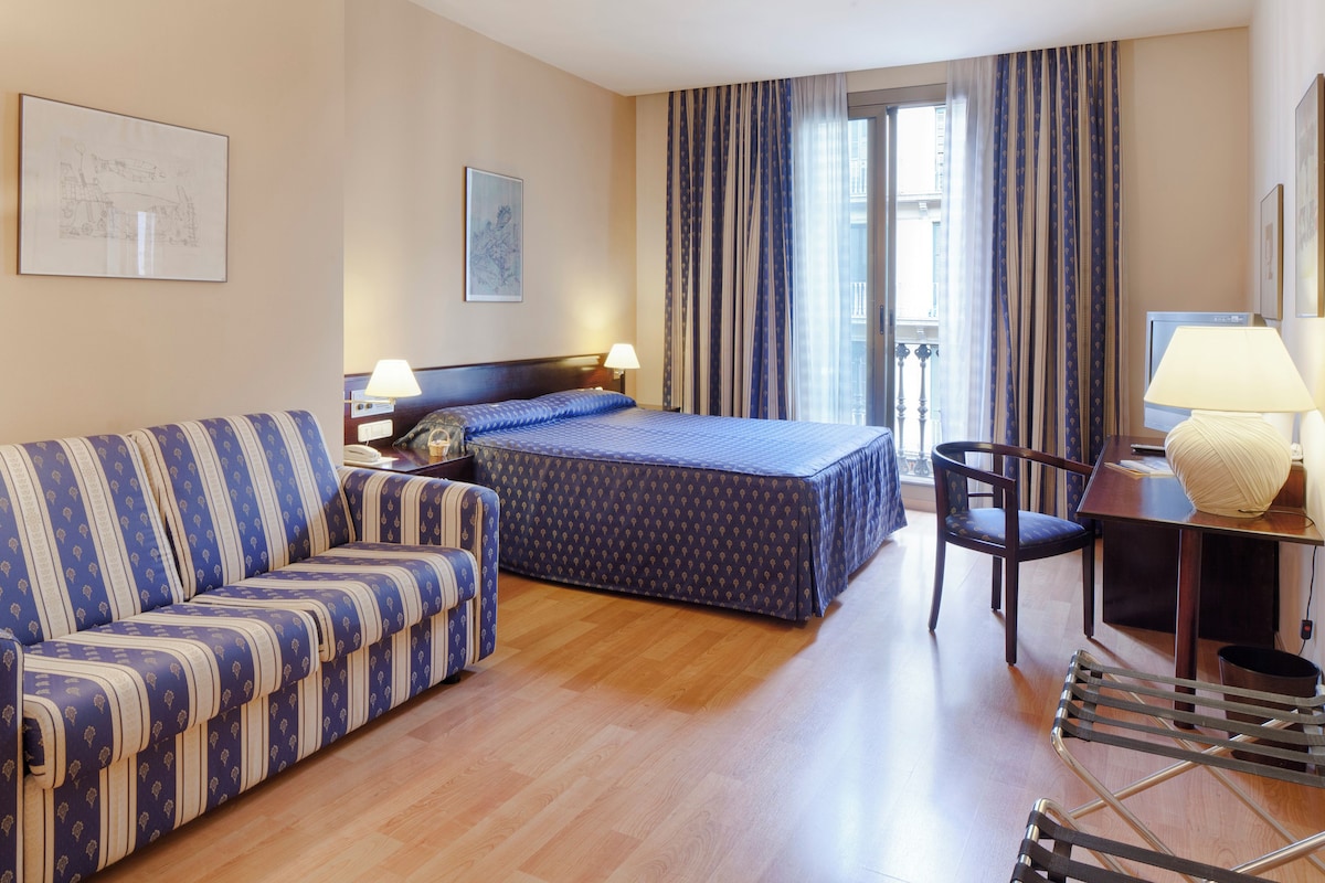 A hotel room with a bed, a sofa, a desk with a lamp, and a chair.