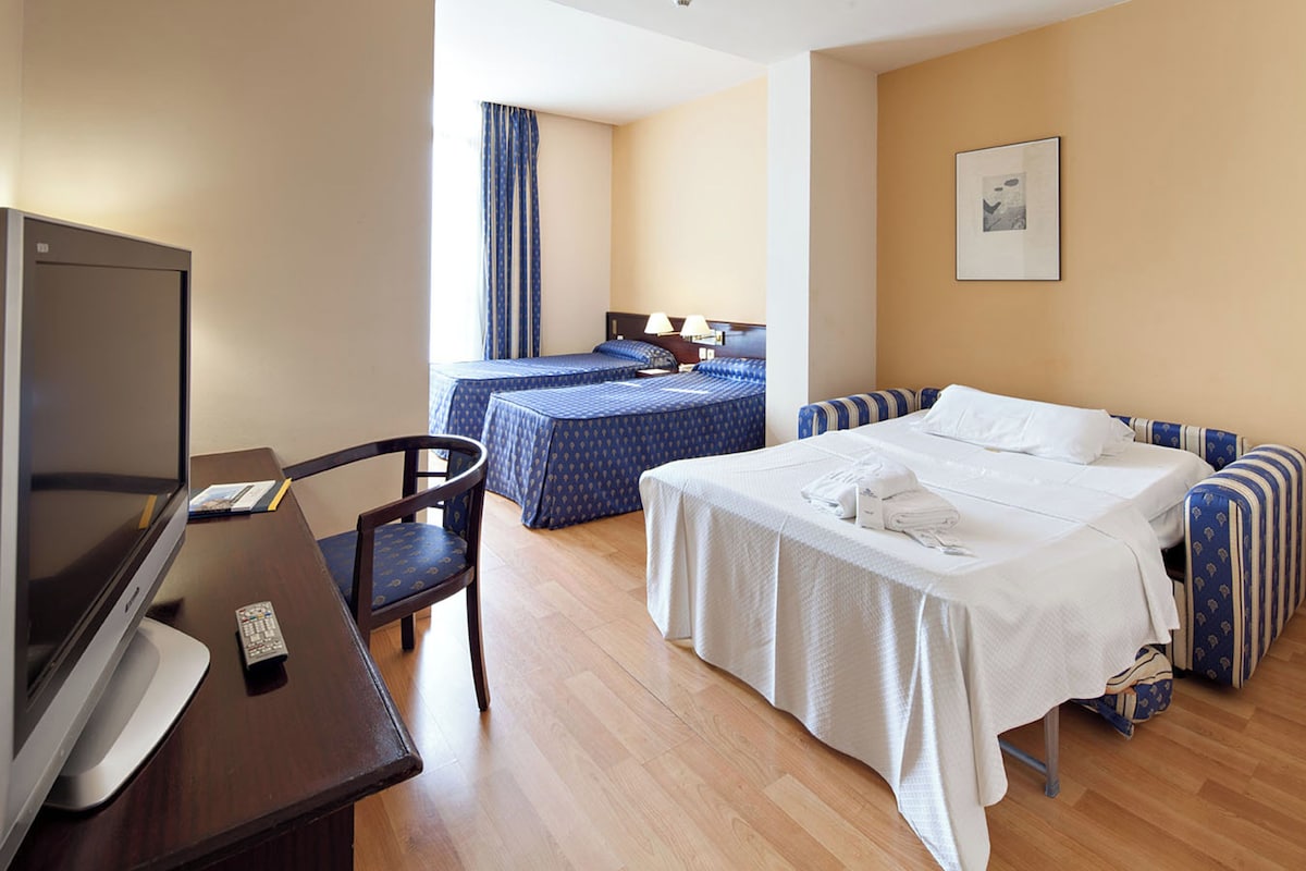 A hotel room with a bed, a desk with a TV, a chair, and a window with curtains.