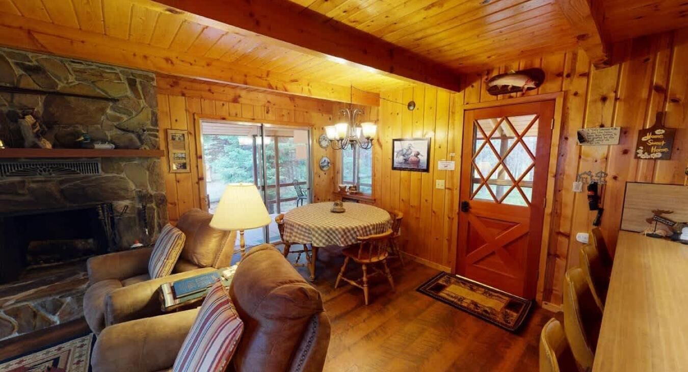 L’intérieur d’un chalet chaleureux, avec une cheminée en pierre, des canapés en cuir, une table à manger et une porte en bois ornée d’une croix.