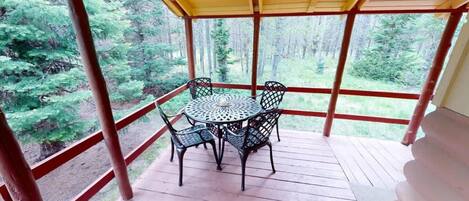 A covered outdoor seating area with a table and chairs, surrounded by a wooden deck and a view of a forested landscape.
