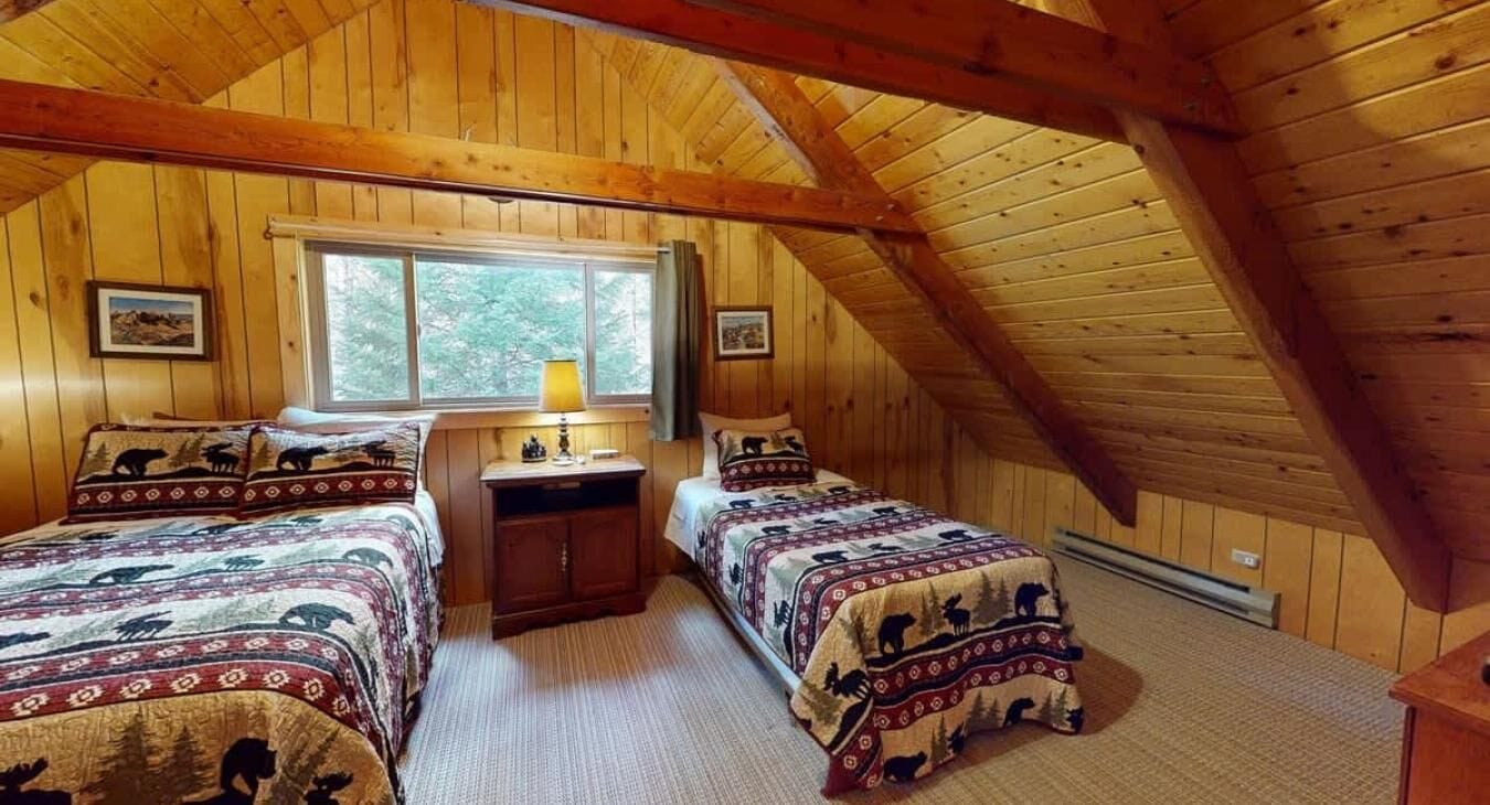 A cozy attic bedroom with wooden walls, two beds with patterned bedding, a window, and a desk.
