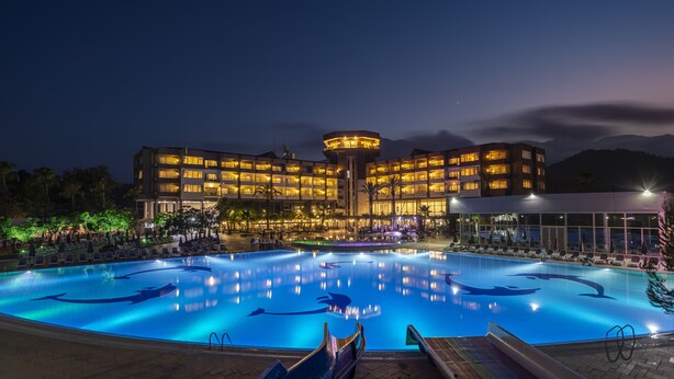 A hotel with a swimming pool featuring dolphin designs, surrounded by a well-lit outdoor area with lounge chairs and a mountain backdrop. - Simena Comfort Hotel (Kemer)