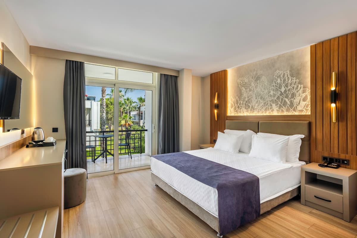 A modern hotel room with a large bed, a desk with a flat-screen TV, and a balcony with outdoor seating.