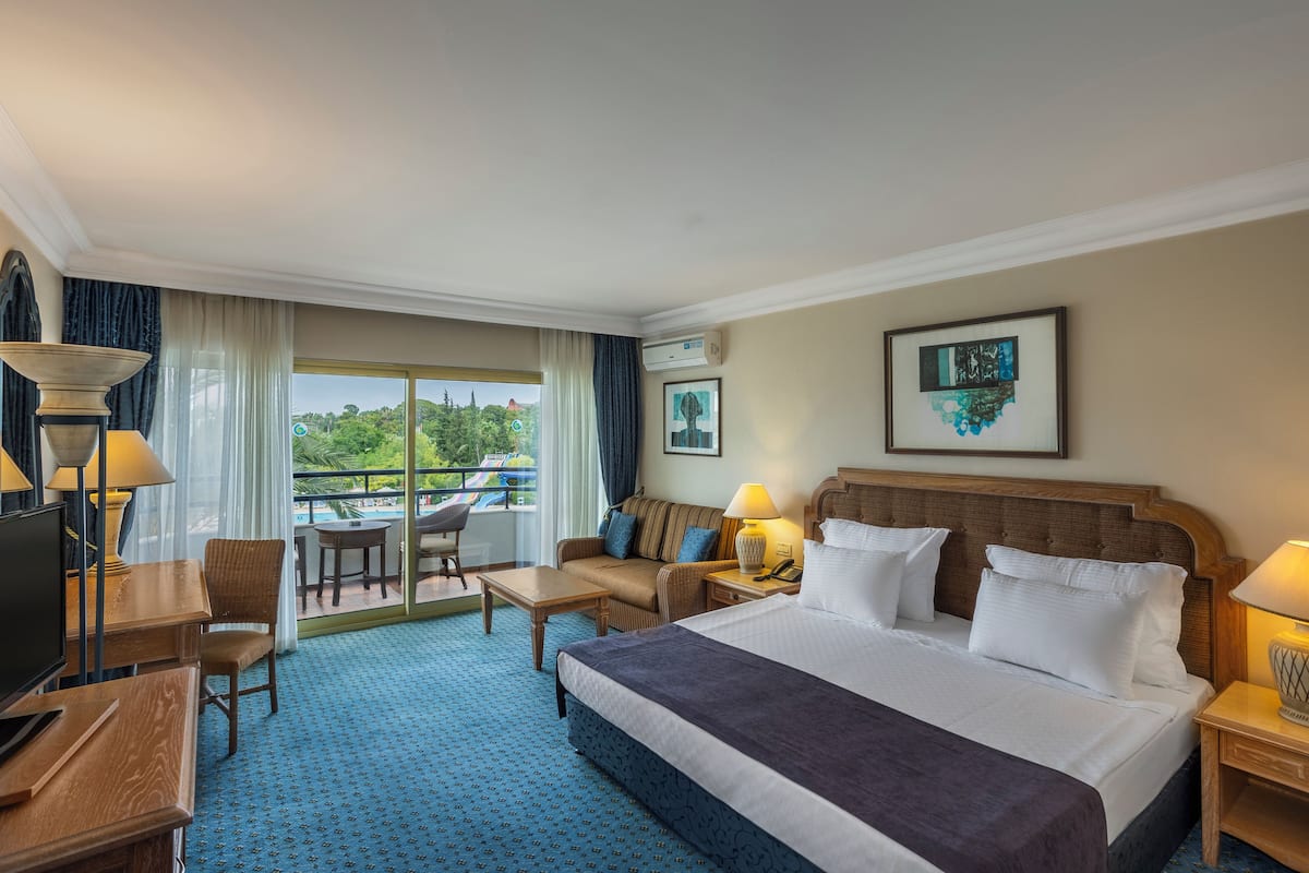 A hotel room with a large bed, a desk, a chair, a sofa, and a balcony with a pool view.