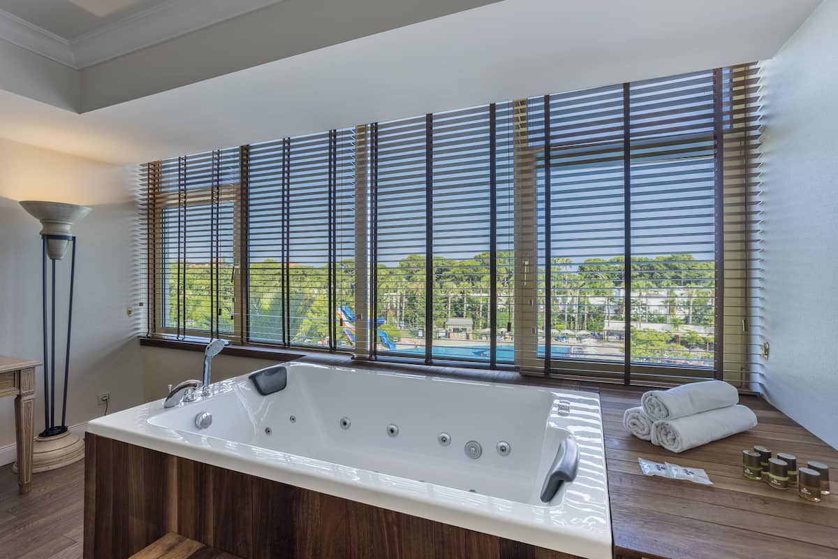 A modern bathroom with a large bathtub, wooden flooring, and a view of a swimming pool through large windows.