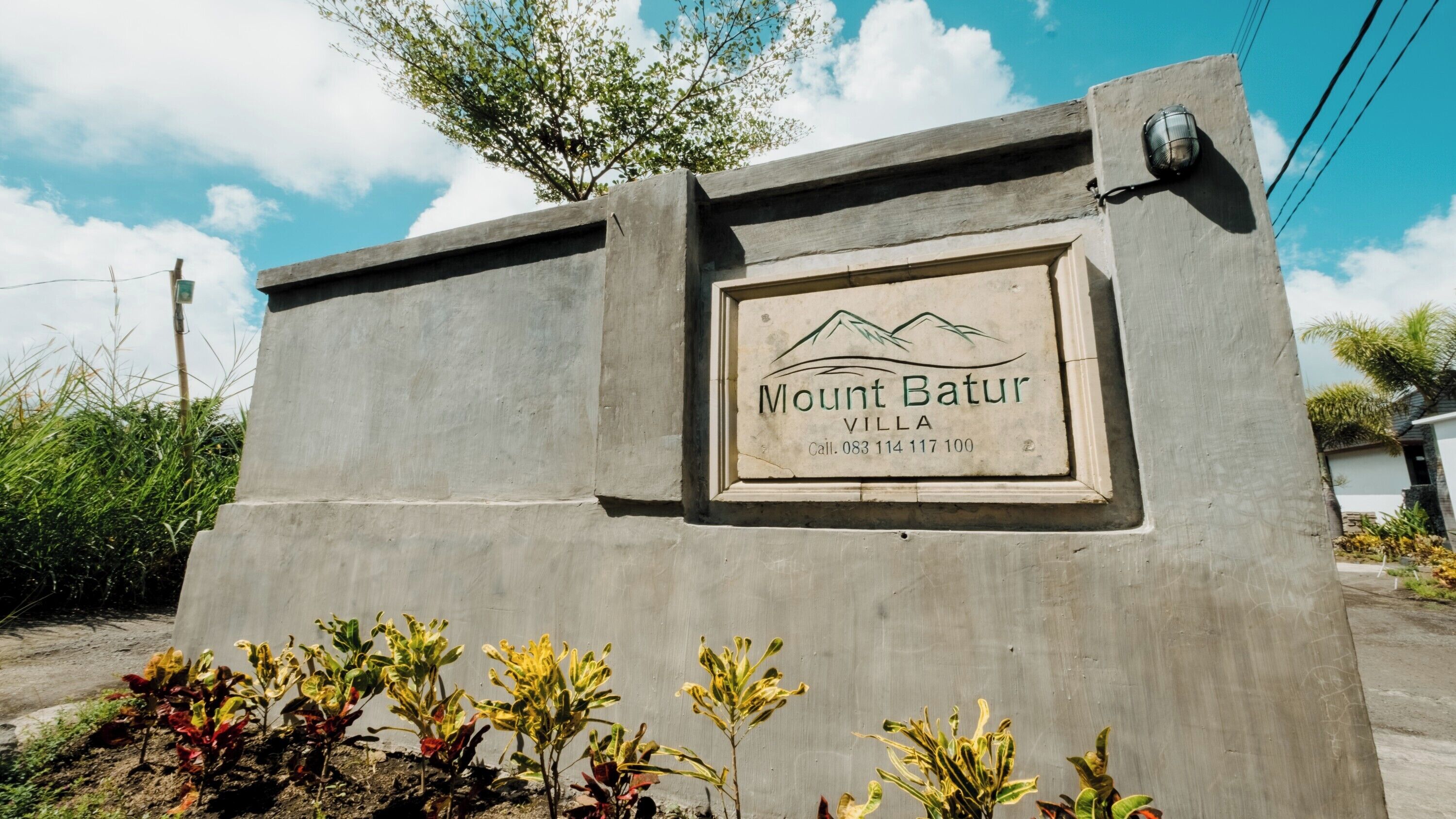 Segnaletica per Mount Batur Villa con grafica raffigurante una montagna, un numero di contatto e una struttura in cemento con piante antistanti.