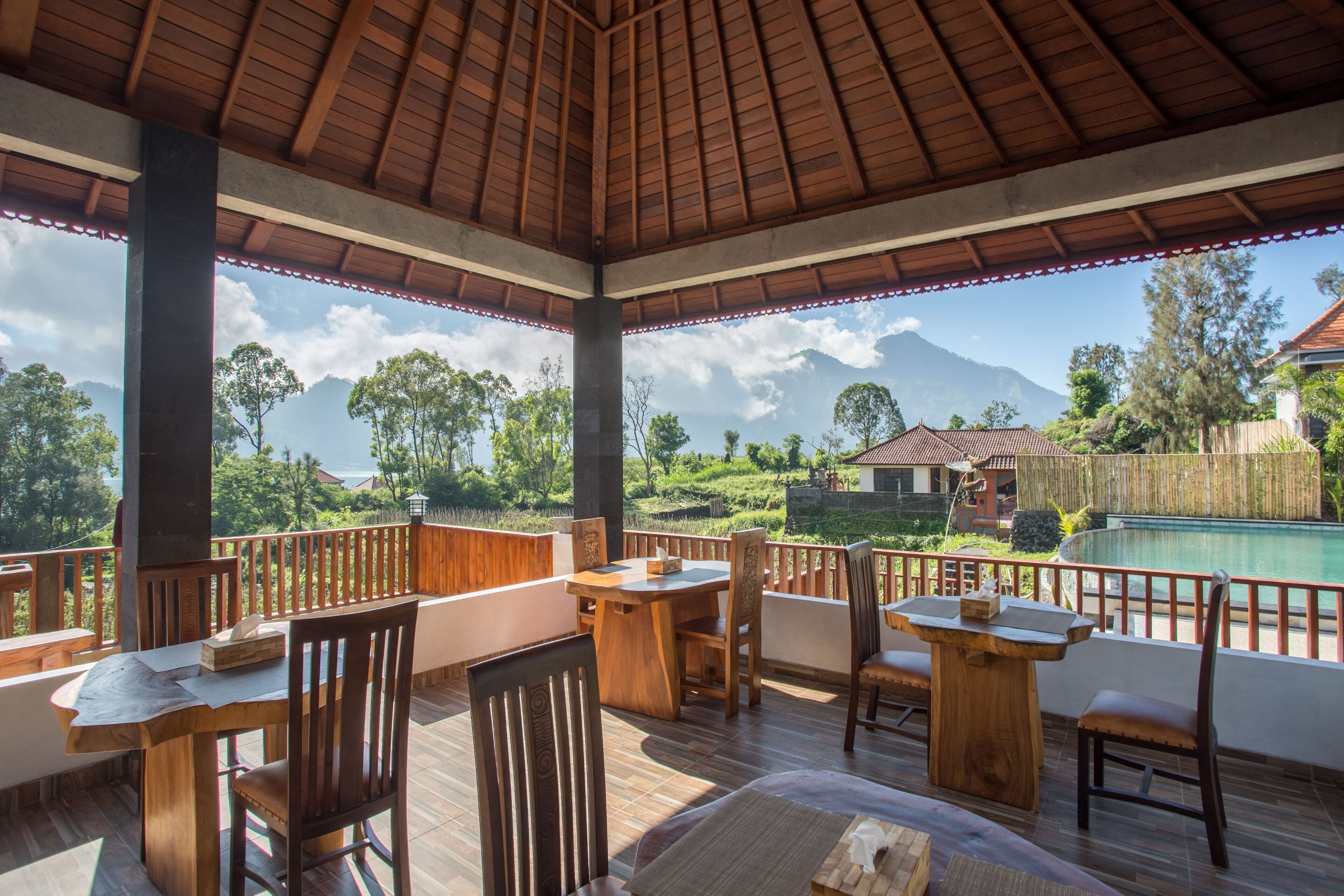 A covered outdoor dining area with wooden furniture and a view of a swimming pool and mountains.