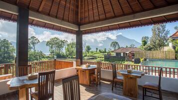 A covered outdoor dining area with wooden furniture and a view of a swimming pool and mountains.
