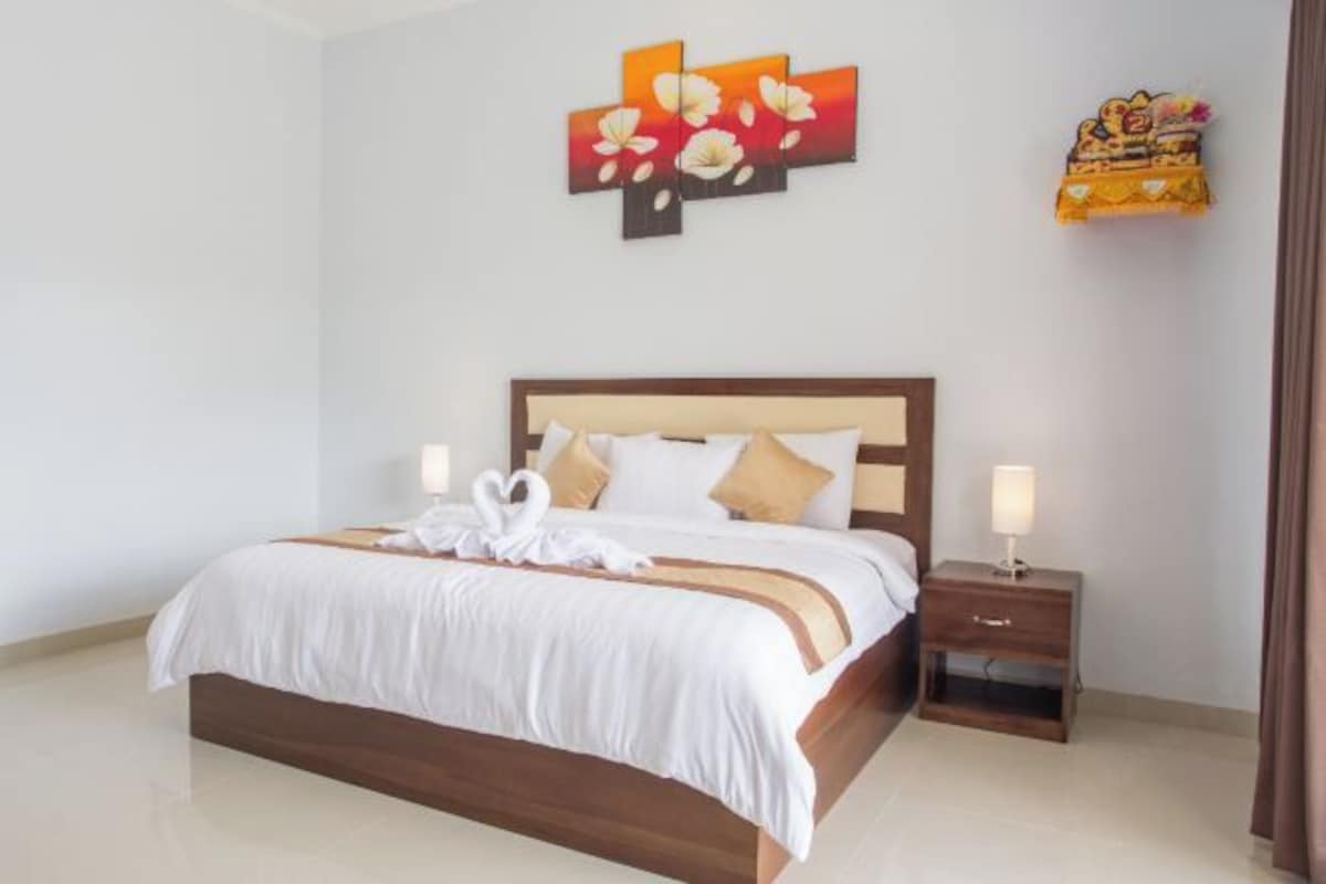 A neatly made bed with white linens and a wooden headboard, a bedside table with a lamp, and a wall with a floral painting.
