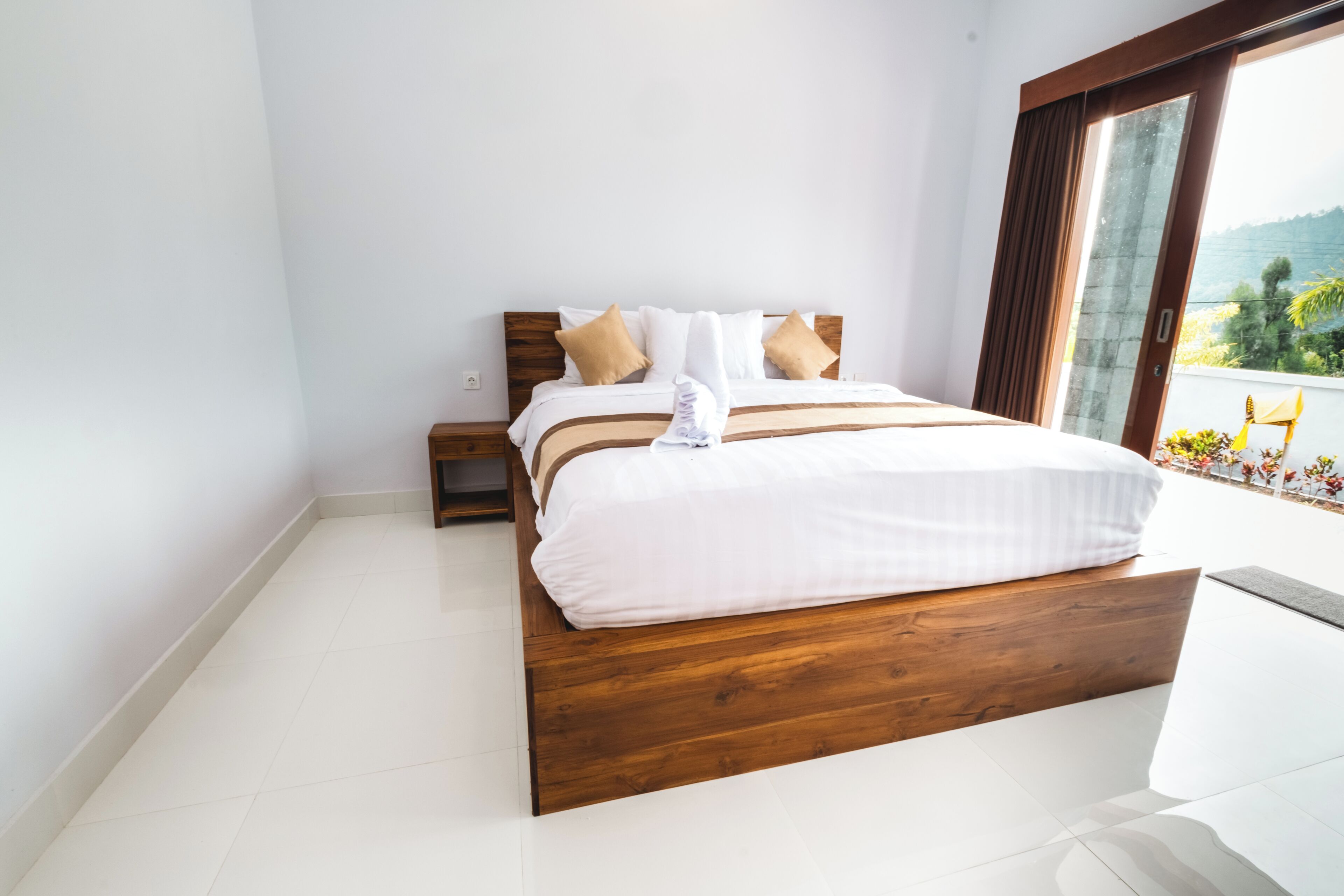 Ein modernes Schlafzimmer mit einem hölzernen Bett, weißer Bettwäsche und Blick auf einen Balkon mit Pflanzen.