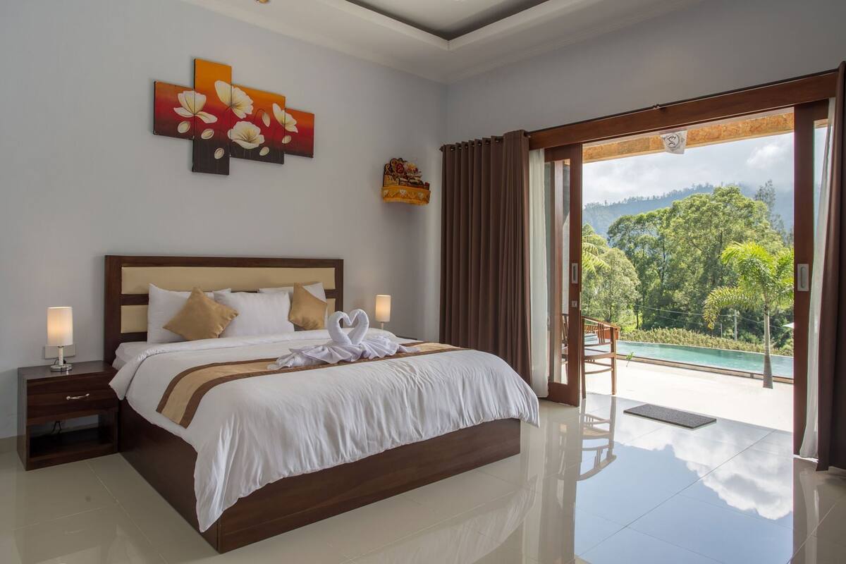 A bedroom with a bed, a nightstand, a lamp, a painting, and a view of a pool and greenery.