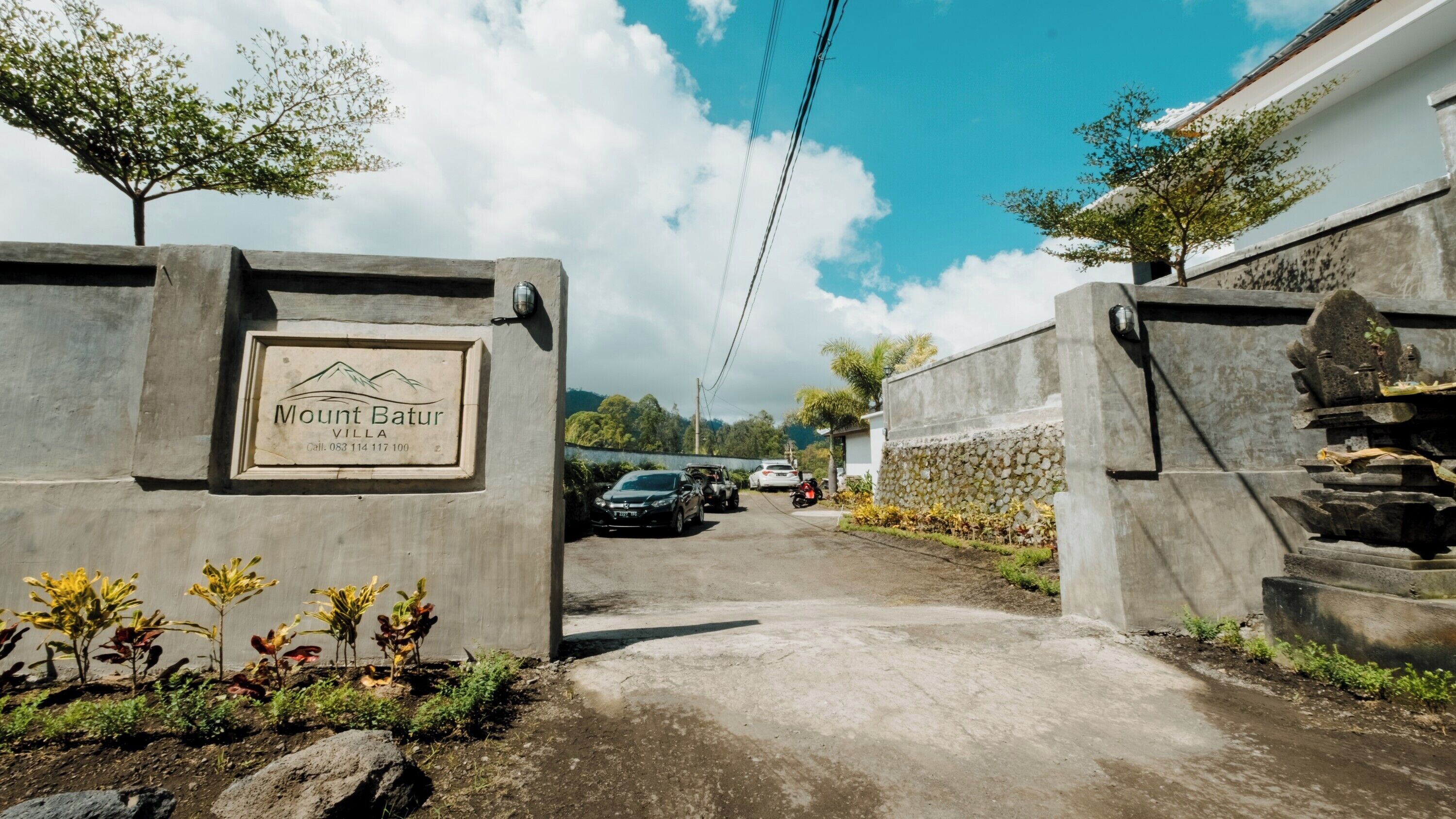 A villa entrance with a sign reading "Mount Batur Villa" and a paved driveway leading to parked vehicles.