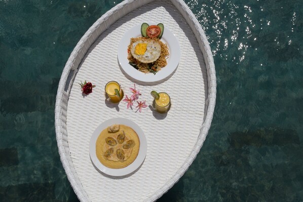 A white wicker boat with a plate of food and drinks on it, floating on clear water.
