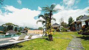 Zona de piscina con sillas de descanso, una palmera y un edificio tradicional al fondo.