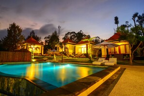 Una piscina con sillas de descanso y sombrillas, rodeada de edificios con techos rojos y un jardín ajardinado.