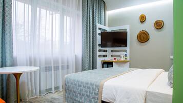 A hotel room with a bed, a TV, a small table, and a window with sheer curtains.