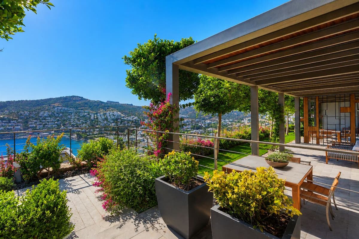 A modern outdoor patio with a view of a coastal city, surrounded by lush greenery and potted plants.