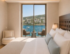 A hotel room with a large bed, two chairs, a small table, and a view of a coastal town and water.