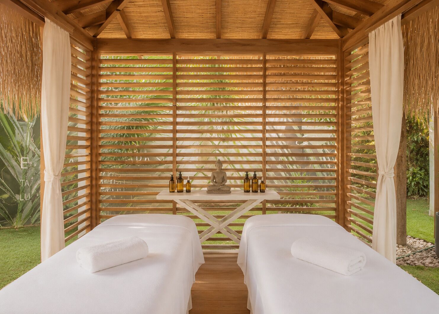A spa room with two massage tables, a wooden ceiling, and a view of greenery.