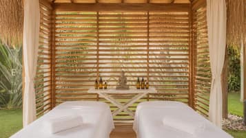 A spa room with two massage tables, a wooden ceiling, and a view of greenery.