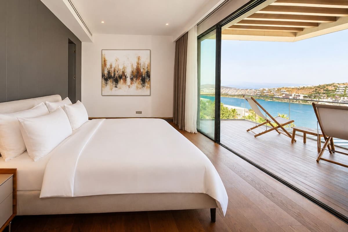 A modern bedroom with a large bed, a bedside table, a nightstand, and a view of a balcony with outdoor seating.