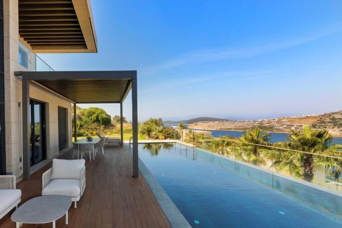 A modern outdoor pool area with a wooden deck, white outdoor furniture, and a view of the sea.