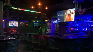 A dimly lit bar area with neon lights, a TV screen displaying a movie, and a bar counter with a drink menu. - Out Back (Olongapo)