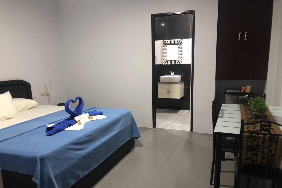 A hotel room with a bed, a desk, a chair, a small table, a plant, a mirror, and a cabinet.