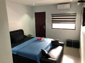 Un dormitorio con cama, ventana con persianas, puerta, equipo de aire acondicionado y una planta pequeña.