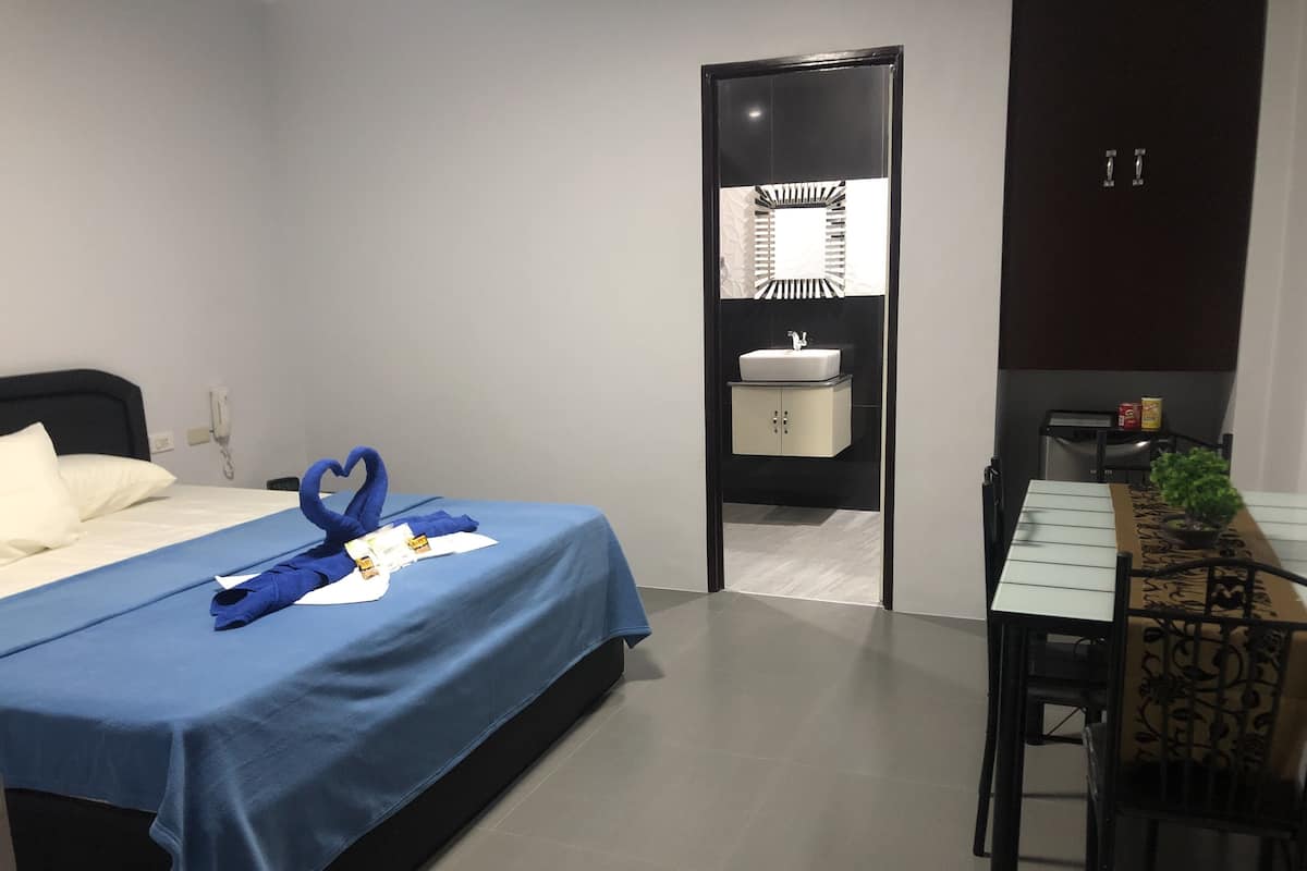 A hotel room with a bed, a desk, a chair, a small table, a plant, a mirror, and a cabinet.