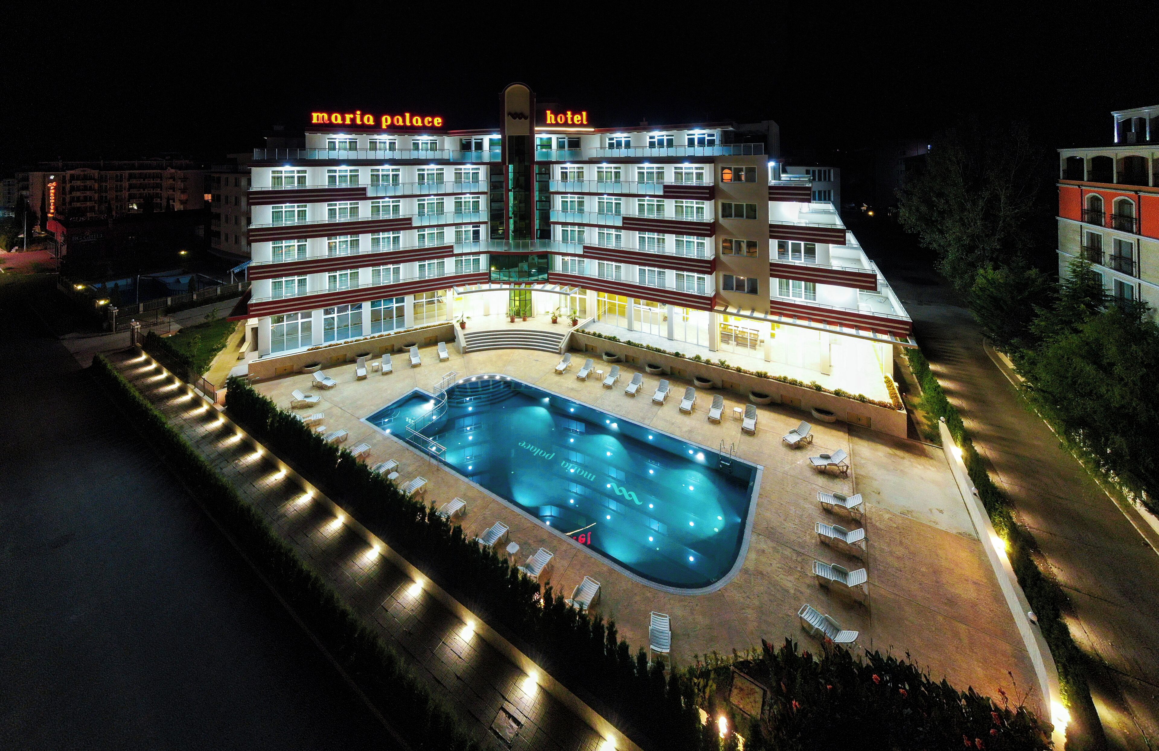 Ett hotell med flera våningar, en swimmingpool, solstolar och en väl upplyst fasad.