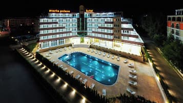 Ett hotell med flera våningar, en swimmingpool, solstolar och en väl upplyst fasad.