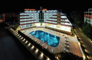 A multi-story hotel with a swimming pool, lounge chairs, and a well-lit exterior.