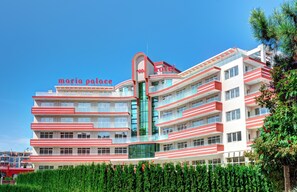 A multi-story hotel with a distinctive red and white facade, balconies, and a central glass elevator shaft. - Maria Palace (Nessebar)