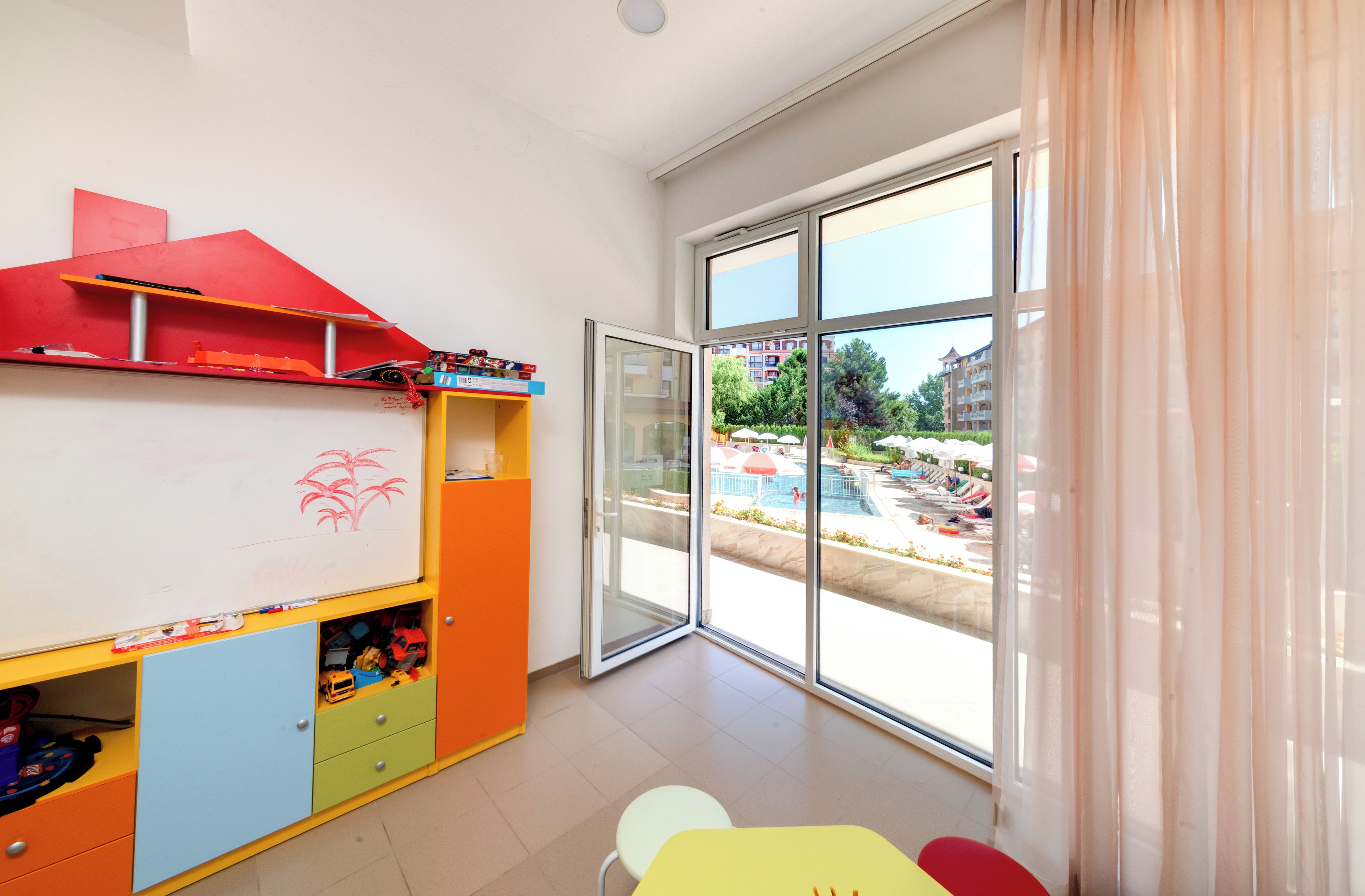 Un área de juegos infantiles con una pizarra, contenedores de almacenamiento coloridos y un amplio ventanal corredera que da a una zona de piscina al aire libre.