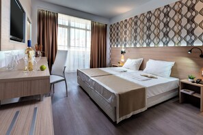 A hotel room with a bed, a desk with a bottle and glasses, a chair, a TV, and a patterned wall. - Maria Palace (Nessebar)