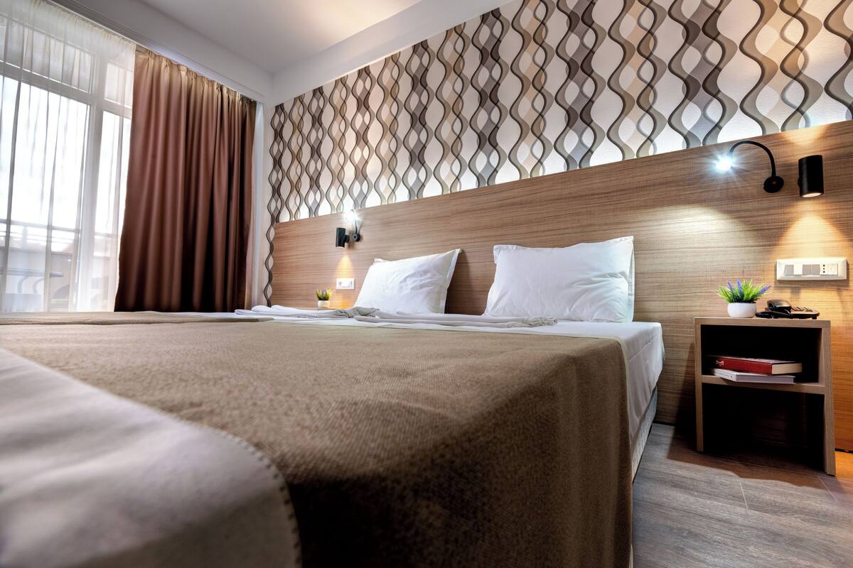 A modern hotel room with a large bed, a wooden headboard, a patterned wall, a bedside table with a lamp, and a small plant.