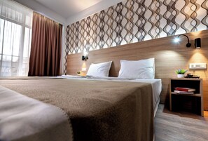 A modern hotel room with a large bed, a wooden headboard, a patterned wall, a bedside table with a lamp, and a small plant. - Maria Palace (Nessebar)